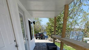 En overdækket terrasse med klinkegulv, en bænk og udsigt til en sø.