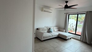 Quarto com sofá branco, ventoinha no teto, ar condicionado e uma janela grande com cortinas.