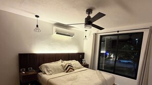 Quarto com cama, mesa de cabeceira, ventilador de teto, ar condicionado e uma janela grande.