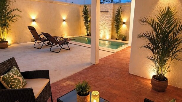 Um pátio externo com móveis de vime, piscina e luzes fixadas na parede.