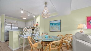 A dining area with a glass table, wicker chairs, a chandelier, and a kitchen with white cabinets and a refrigerator. - Island Beach and Racquet Club B104: Oceanfront Condo with Views, Community Amenities (Atlantic Beach)
