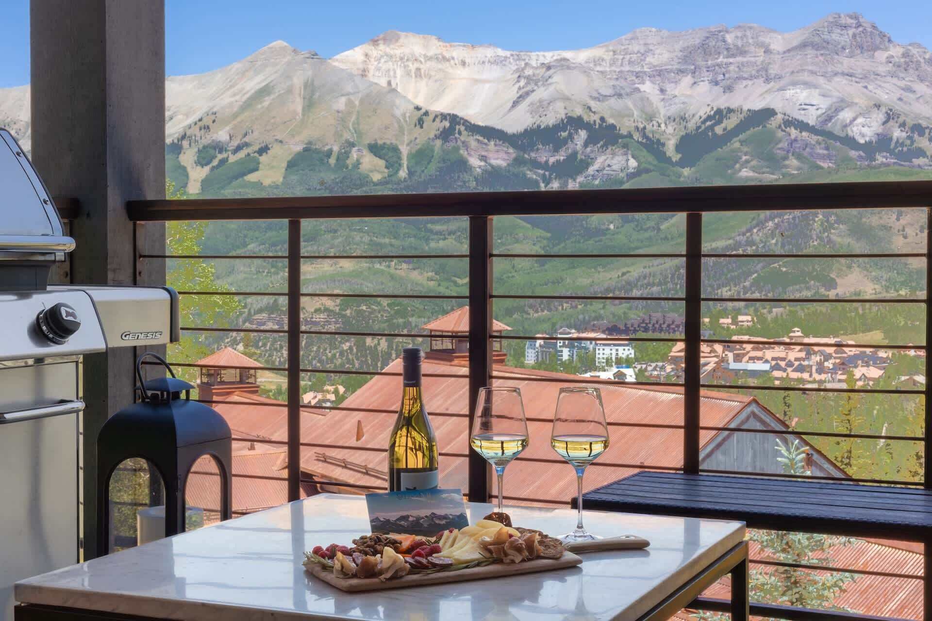 A balcony with a view of mountains, a grill, a bottle of wine, and a cheese board.