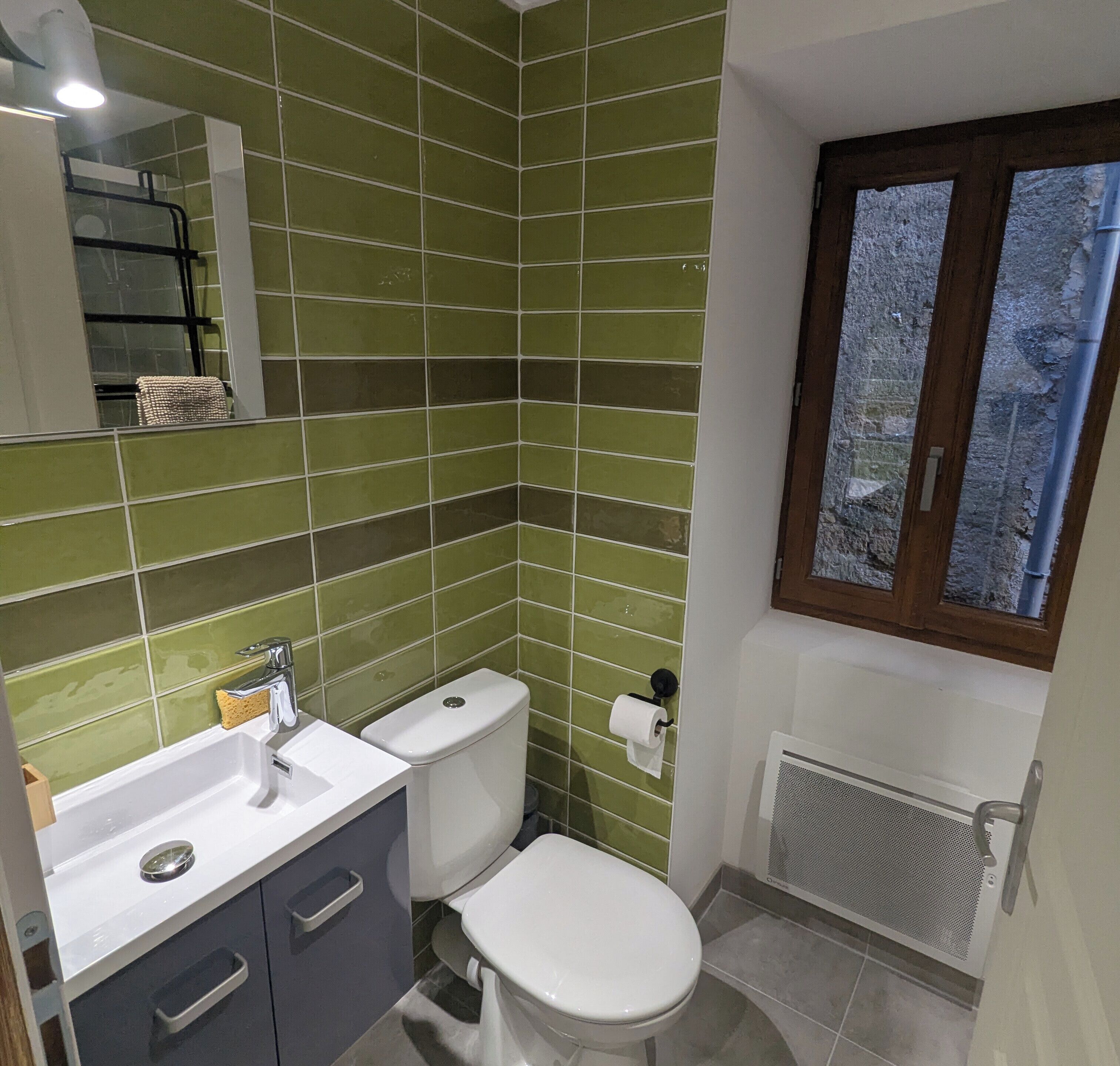 A bathroom with green tiled walls, a white sink, a toilet, and a window with a view.