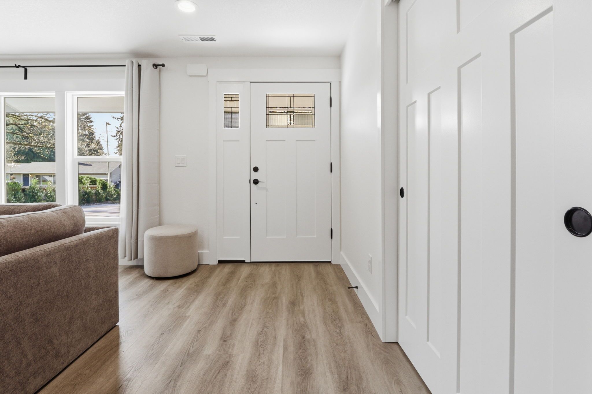 A room with a wooden floor, a beige sofa, a white door with a window, and a small round ottoman.