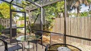 Un patio cubierto de vidrio con una mesa y sillas, rodeado por una cerca de bambú y follaje tropical.