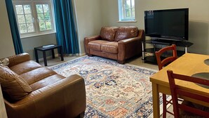 A living room with a brown leather sofa, a wooden dining table, a TV, and a patterned rug.