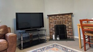 A living room with a flat-screen TV on a stand, a brick fireplace, a leather armchair, and a wooden dining table with chairs.