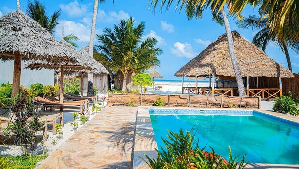A pool area with thatched-roof structures, palm trees, and a clear blue sky. - Zanzibar holiday villa (Zanzibar)