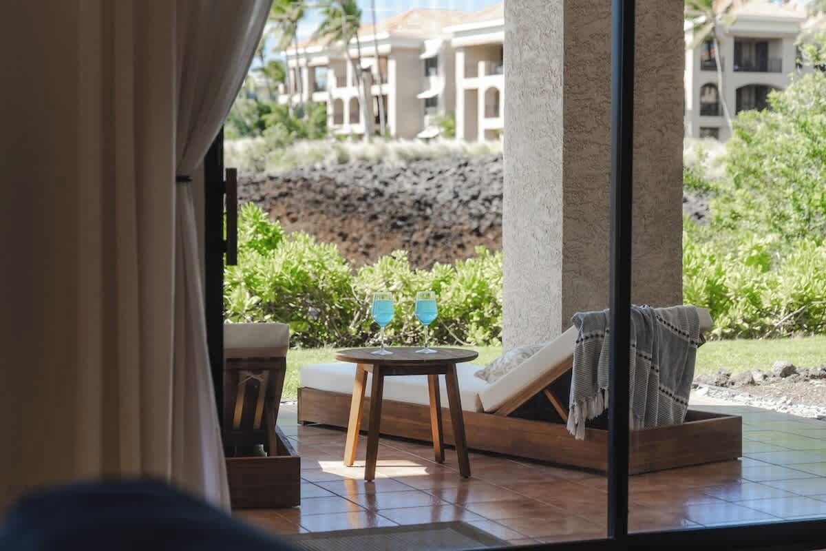 A sun lounger, a small round table with two glasses, and a view of a resort area with buildings and greenery.