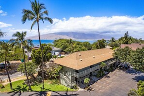 A hotel with a parking lot, a building with a flat roof, and a view of the ocean and mountains. - Ocean View Escape | Lanai, Pool & Maui Sunsets (Kihei)