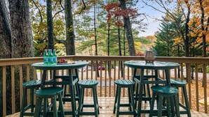 Une terrasse en bois, avec des chaises et des tables vertes, donnant sur une zone boisée aux couleurs automnales.
