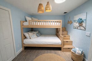Ein Schlafraum mit Etagenbetten, einer Holzleiter, zwei Hängelampen, einem an der Wand befestigten Bild und einem Korbwäschesammler mit Handtüchern.