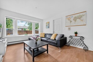 A modern living room with a black leather sofa, a coffee table, and a world map on the wall. - Spacious 2BR Ground-Level Suite Near Kingsway Central Park & Metrotown 🌿 (Vancouver)