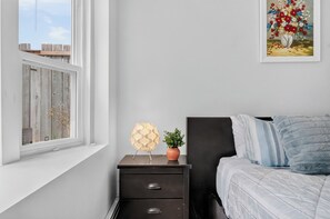 Et soveværelse med et vindue, et natbord med en lampe, en potteplante og en seng med puder.