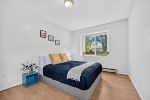 A bedroom with a bed, a window, a wall-mounted light, and a small blue nightstand with a vase. - Spacious 2BR Ground-Level Suite Near Kingsway Central Park & Metrotown 🌿 (Vancouver)