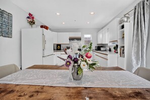 A modern kitchen with white cabinets, a wooden dining table, and a vase of flowers. - Spacious 2BR Ground-Level Suite Near Kingsway Central Park & Metrotown 🌿 (Vancouver)