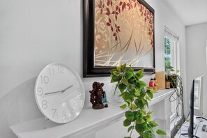En hvid væg med et ur, et blomsterbillede i ramme, en reol med figurer og en plante.