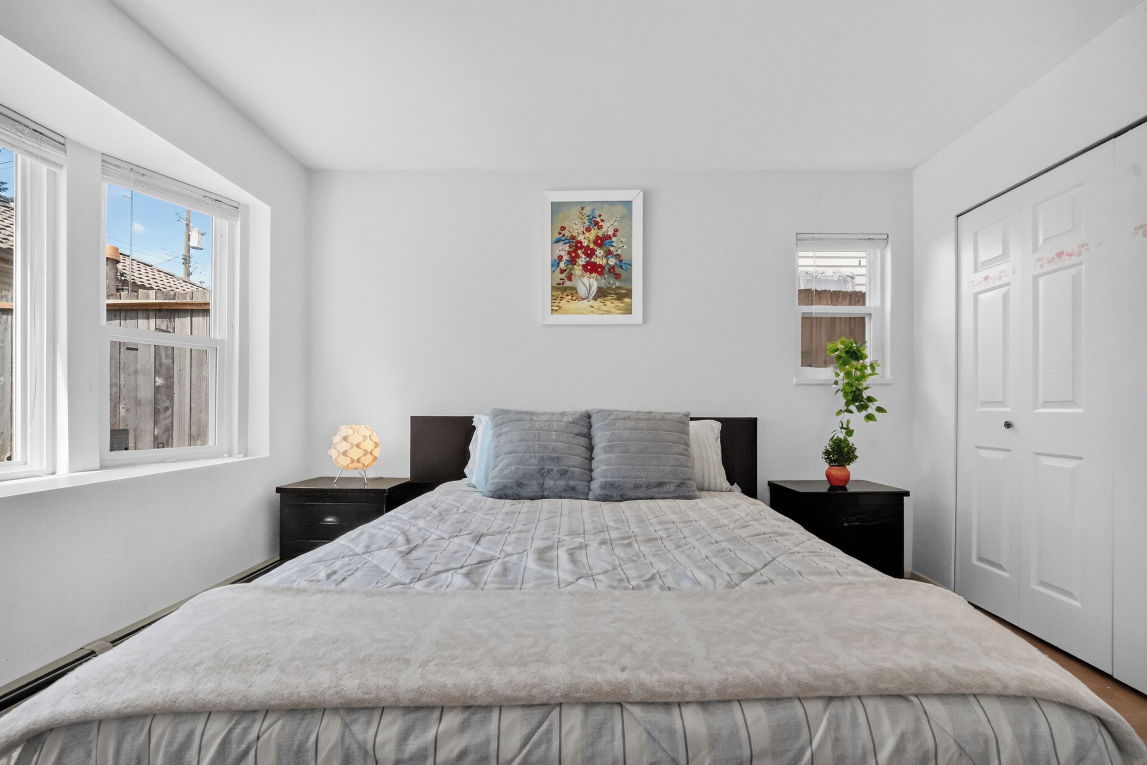 Une chambre à coucher avec un lit, deux tables de chevet, une commode, une plante en pot, un tableau et une fenêtre.