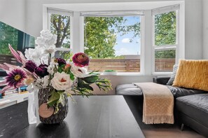 A room with a view of trees through large windows, a black leather sofa, and a vase of flowers on a dark table. - Spacious 2BR Ground-Level Suite Near Kingsway Central Park & Metrotown 🌿 (Vancouver)