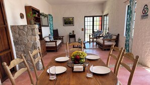 A dining area with a wooden table, chairs, a fruit bowl, and a stone wall.