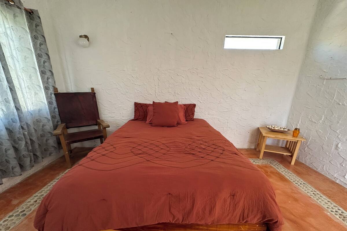 A bedroom with a wooden bed, a chair, a small table with a bowl and jar, and a window.