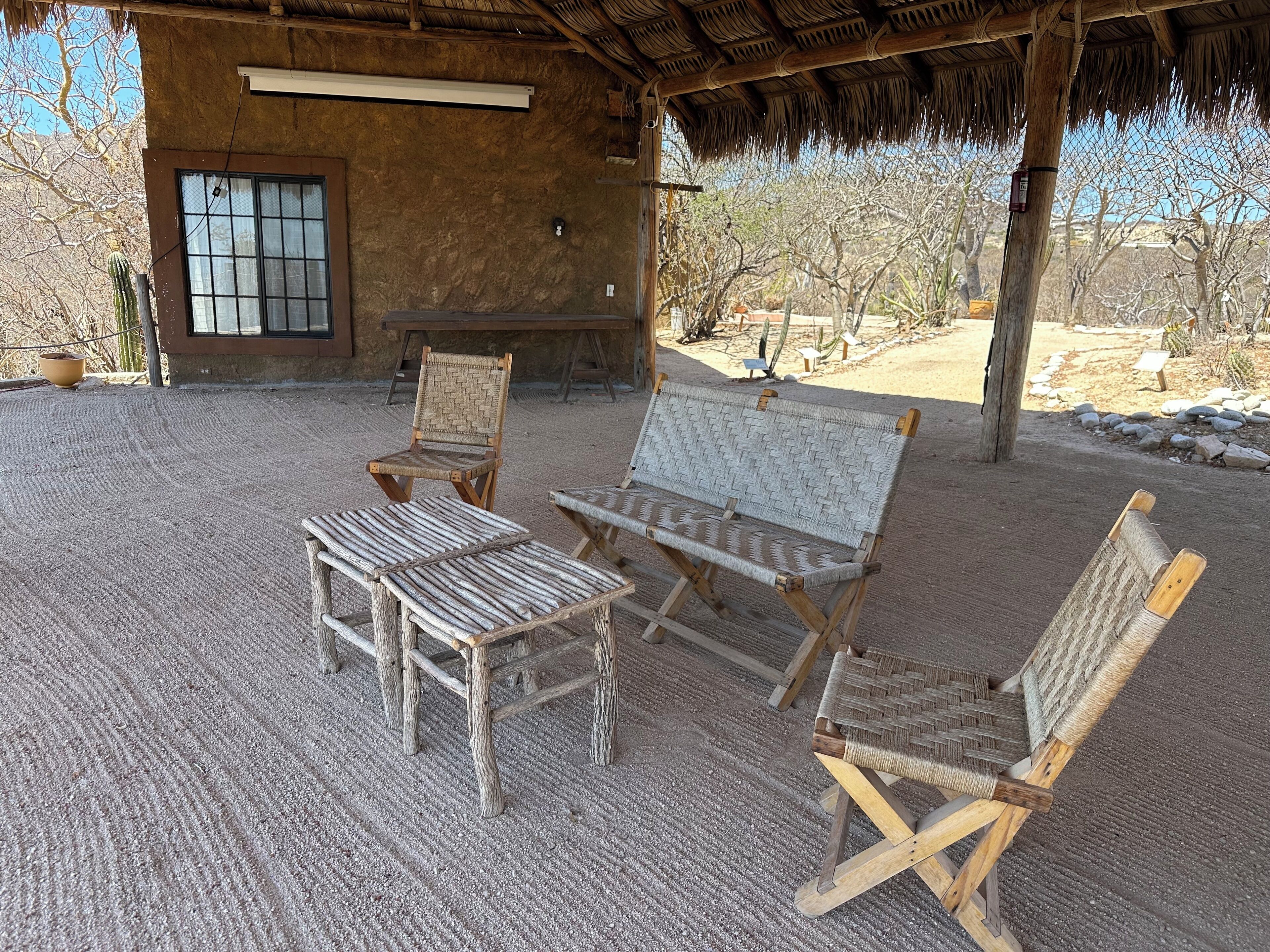 Un área de estar al aire libre rústica con sillas tejidas y una mesa, una estructura cubierta de paja y una ventana con persianas.