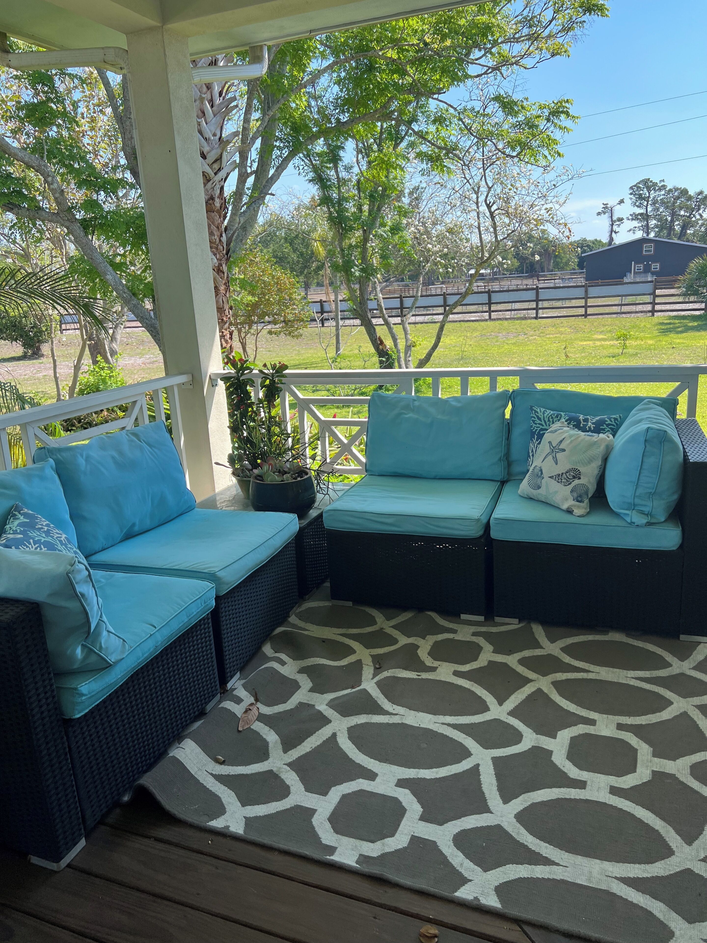A covered outdoor seating area with wicker furniture and blue cushions, a patterned rug, and a view of a fenced yard with trees and a building in the background.