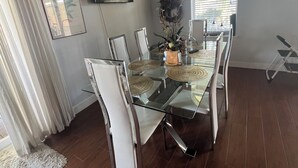 Zona pranzo con tavolo in vetro, sedie bianche, ventilatore a soffitto, televisore e finestra con persiane.