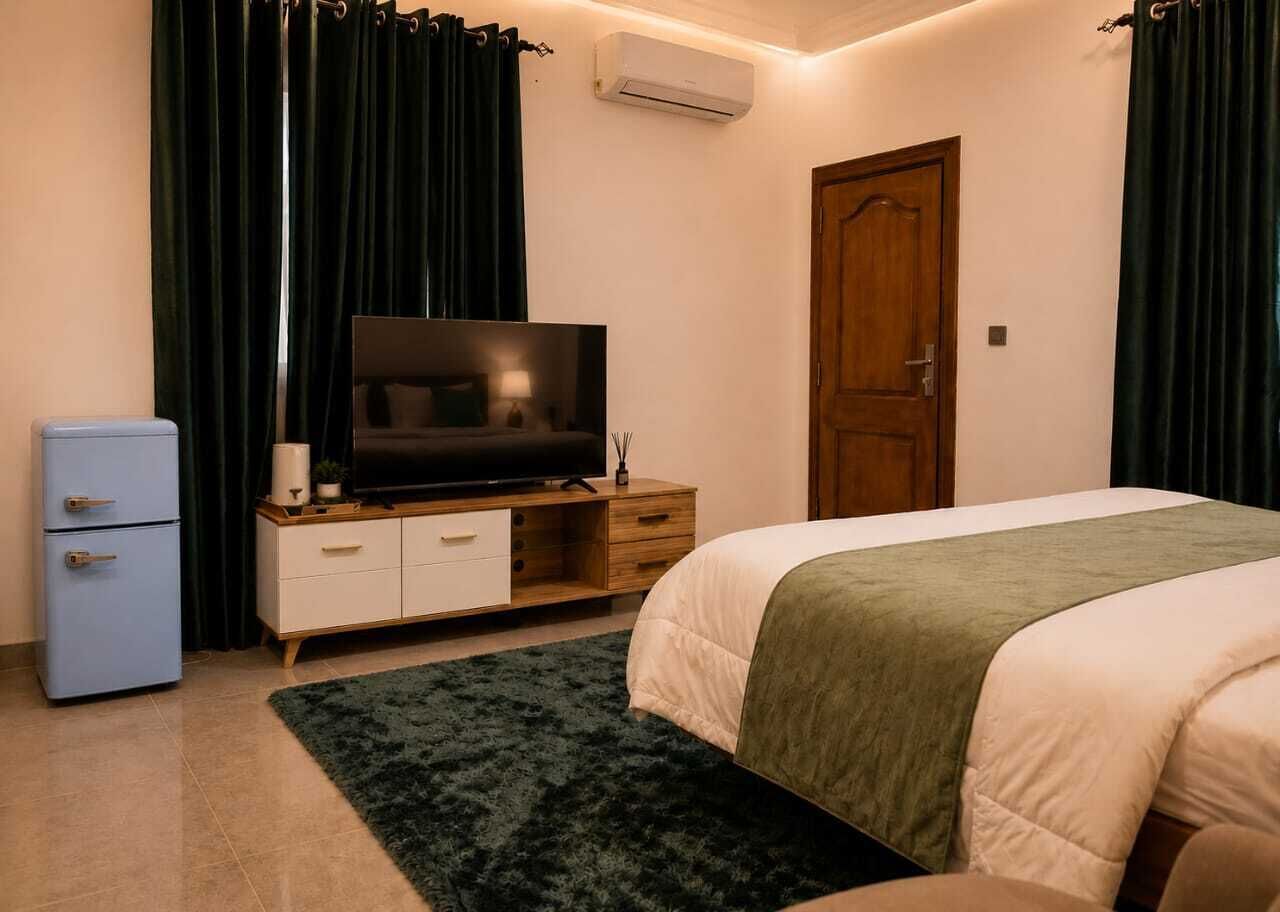 A bedroom with a bed, a TV on a wooden cabinet, a small fridge, and a door.