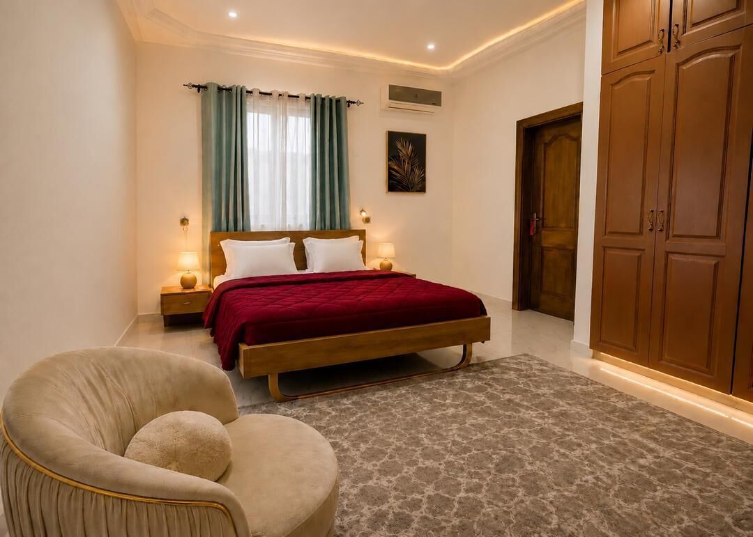A bedroom with a bed, bedside lamps, a chair, a wardrobe, and a window with curtains.