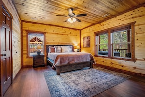 A wooden cabin bedroom with a bed, nightstands, a ceiling fan, and large windows.
