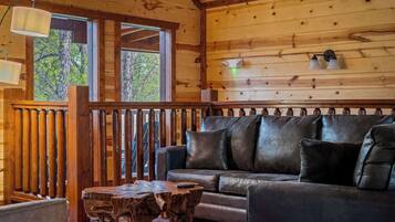 A wooden cabin interior with a leather sofa, a wooden coffee table, and a view of the outdoors through large windows.