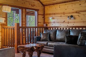 A wooden cabin interior with a leather sofa, a wooden coffee table, and a view of the outdoors through large windows.