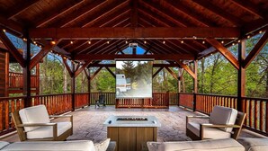 Une terrasse extérieure couverte, aménagée avec des meubles en bois, un foyer et offrant une vue sur un véhicule au loin.
