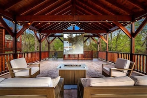 An outdoor covered patio with wooden furniture, a fire pit, and a view of a vehicle in the background. - 8BR Log Cabin | Indoor Pool + Outdoor Theater (Sevierville)