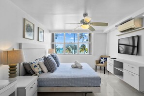 Ein modernes Schlafzimmer mit Bett, Deckenventilator, Fernseher und Fenster mit Blick auf Palmen und den Ozean.