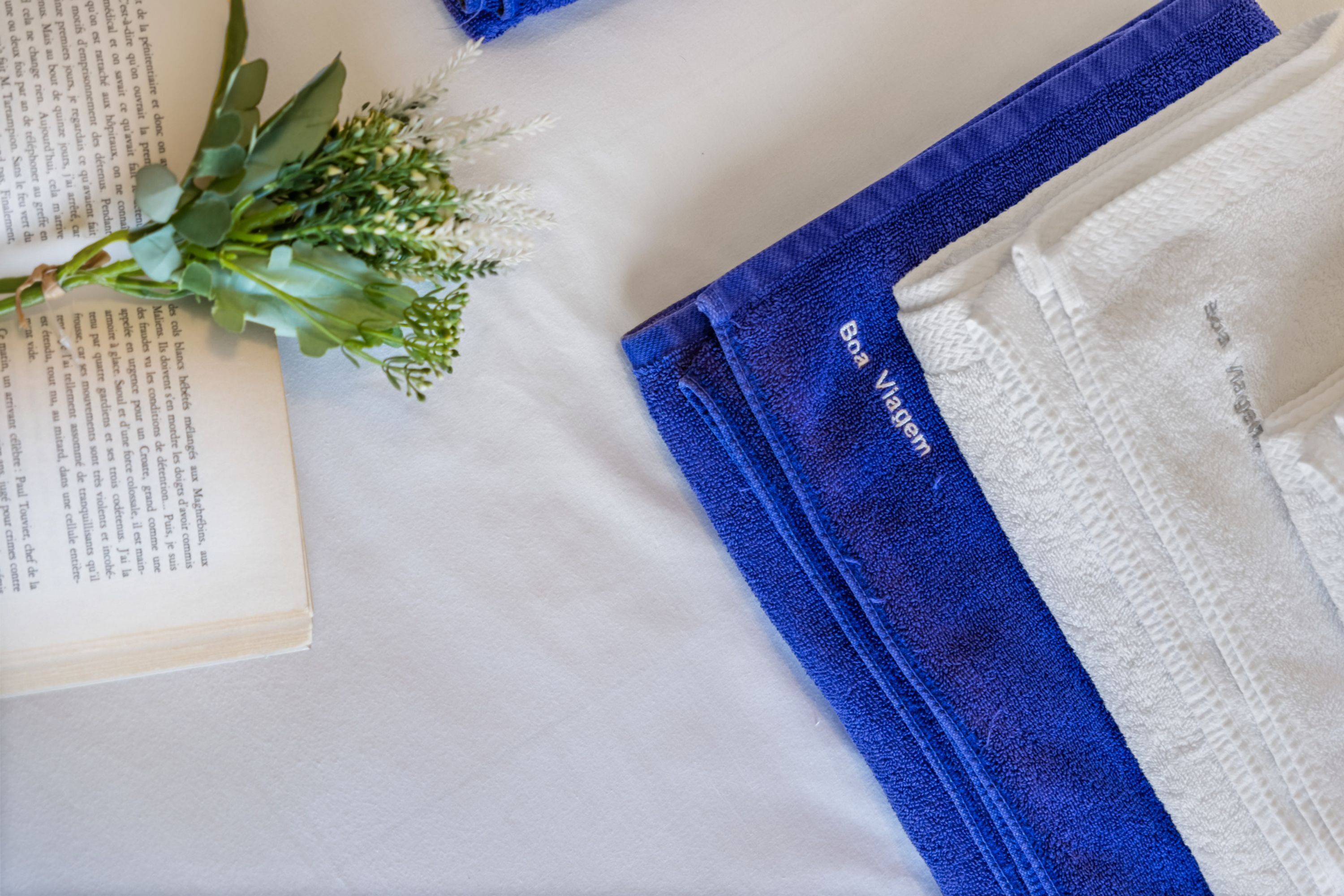 Un lit avec un drap blanc et une couverture bleue, un livre et une petite plante.