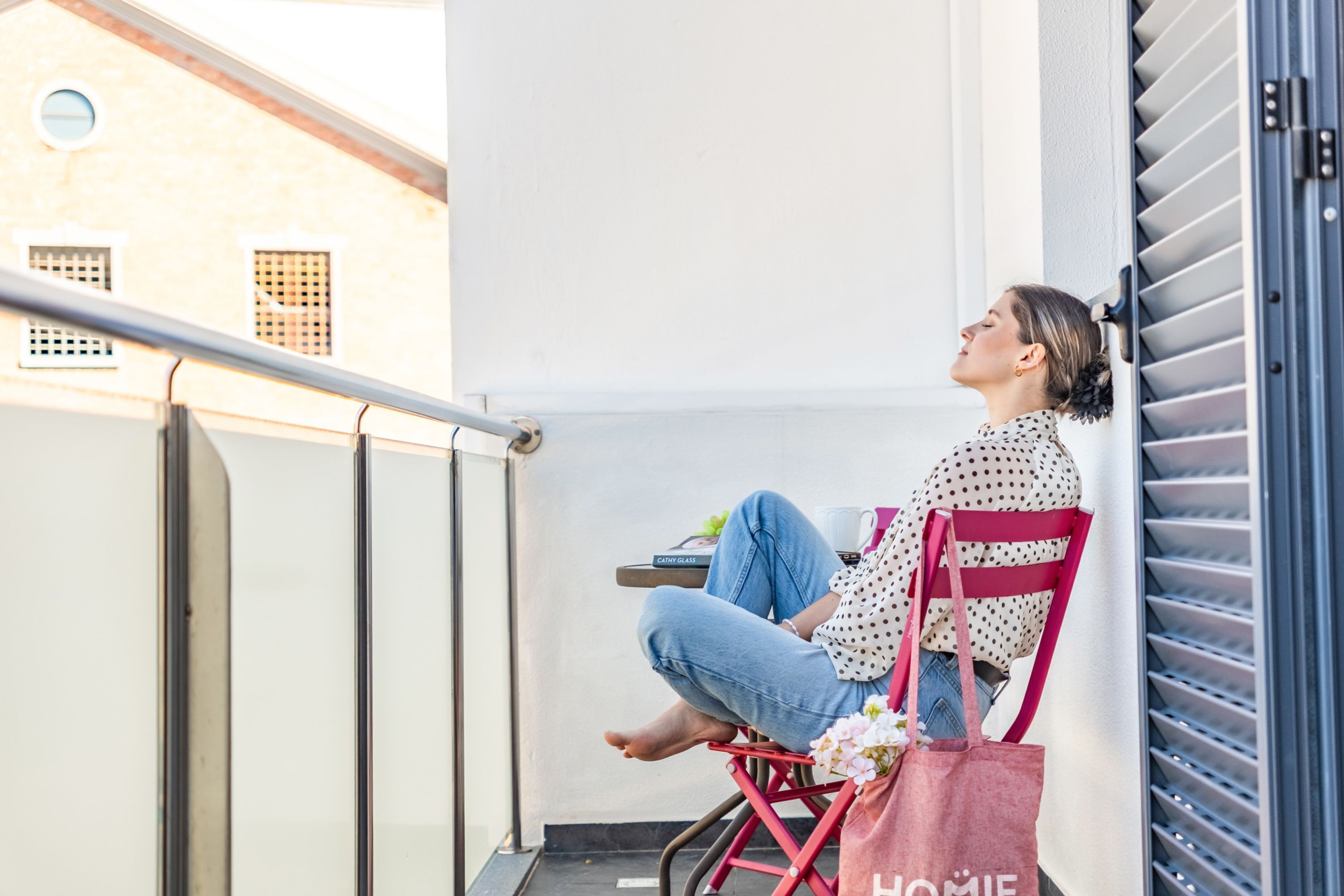 A person sitting on a chair on a balcony with a railing, a building in the background, and a door with horizontal slats.