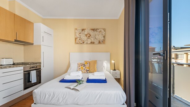 Ett modernt hotellrum med ett kök, en stor säng med handdukar, ett nattduksbord och utsikt genom en skjutdörr i glas.