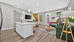 Eine moderne Küche mit einer zentralen Kochinsel, einem Essbereich mit Tisch und Stühlen, einem grünen Sofa und einem an der Wand montierten Fernseher.