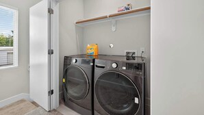 A laundry room with two front-loading washing machines, a shelf with cleaning supplies, and a door leading to another room.
