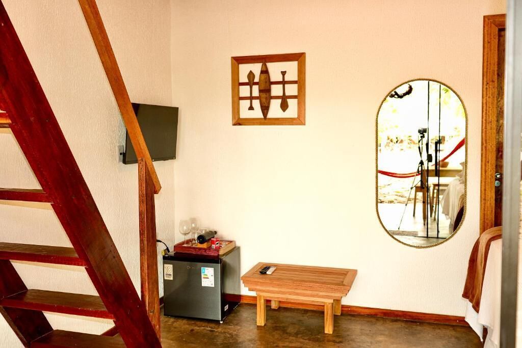 A room with a wooden table, a small refrigerator, a mirror, and a wooden staircase.