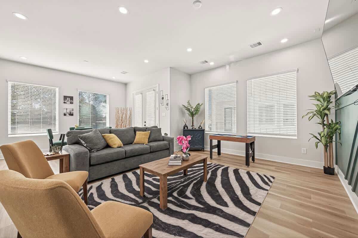 A modern living room with a sofa, coffee table, and chairs, a zebra-patterned rug, and large windows with blinds.
