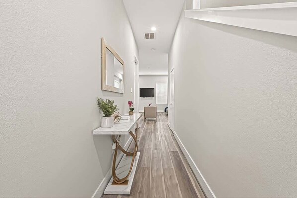 A narrow hallway with a white wall, a mirror, a small table with a vase and flowers, and a wooden floor.