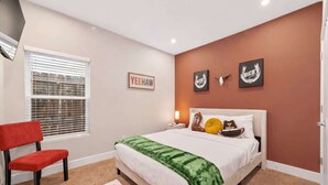 A bedroom with a bed, a red chair, a window with blinds, and wall art. - Texas-Style 3BR Houston + Games (Houston)