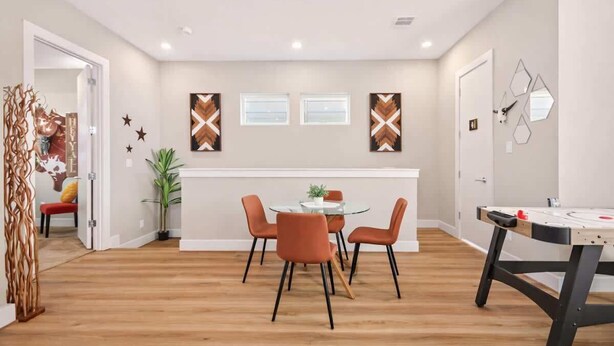 A modern dining area with a round glass table, four orange chairs, and a wall-mounted abstract art piece. - Texas-Style 3BR Houston + Games (Houston)