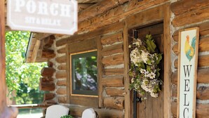 Une cabane en bois avec un porche, une chaise berçante, une pancarte de bienvenue et une porte ornée d’une guirlande.