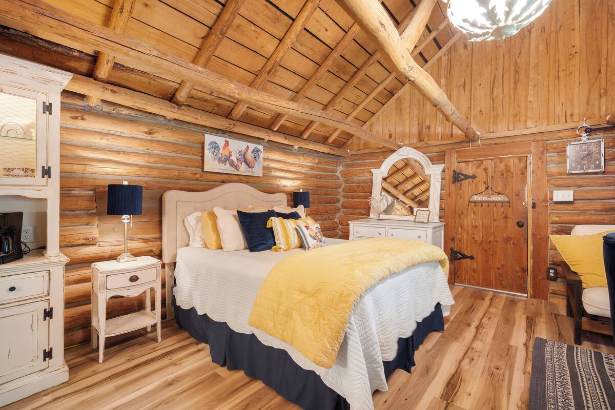 Un dormitorio de estilo cabaña de troncos con cama, mesitas de noche, cómoda y techo de madera.