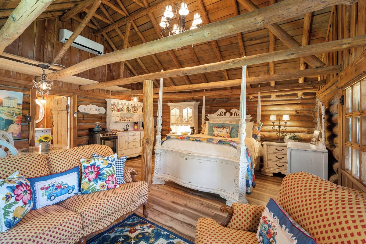 Interior de una cabaña de troncos con una cama, dos sofás, una araña y un comedor.