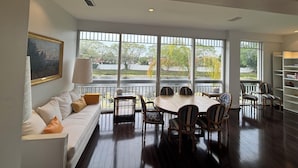 A spacious living area with a dining table surrounded by chairs, a sofa with cushions, a painting on the wall, and large windows offering a view of the outdoors. - Spacious 4-bedroom apartment At IMG Academy (Bradenton)